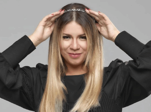 Mulher de cabelos lisos e loiros posa sorrindo levemente diante de um fundo cinza. Ela usa uma blusa preta de mangas bufantes e segura uma tiara prateada sobre a cabeça com as duas mãos.