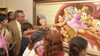 Homem de terno bege e gravata clara, cercado por várias crianças, observa e interage com uma pintura colorida emoldurada. A obra é uma releitura bem-humorada da criação de Adão, de Michelangelo, com personagens infantis de desenho animado no lugar das figuras originais. Algumas crianças apontam para o quadro e uma delas segura um boneco. A cena transmite um ambiente educativo e alegre em uma exposição de arte.