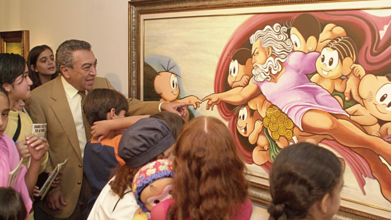 Homem de terno bege e gravata clara, cercado por várias crianças, observa e interage com uma pintura colorida emoldurada. A obra é uma releitura bem-humorada da criação de Adão, de Michelangelo, com personagens infantis de desenho animado no lugar das figuras originais. Algumas crianças apontam para o quadro e uma delas segura um boneco. A cena transmite um ambiente educativo e alegre em uma exposição de arte.