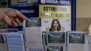 Pessoa segura um volante da “Mega da Virada” em uma casa lotérica. Ao fundo, um cartaz da Mega-Sena exibe a estimativa de prêmio de 600 milhões de reais e a imagem de uma mulher sorridente comemorando.