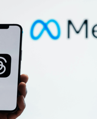 Uma pessoa segura um smartphone que exibe o logotipo do aplicativo Threads, da Meta, em fundo branco. Ao fundo, desfocado, aparece o logotipo da empresa Meta, com o símbolo azul em forma de infinito ao lado da palavra “Meta” em letras pretas. A imagem sugere o vínculo entre o aplicativo e a empresa controladora das redes sociais Facebook e Instagram.