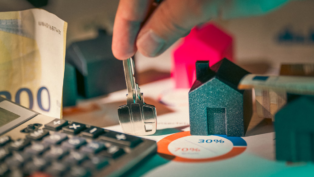 Pessoa segura uma chave sobre uma mesa com miniaturas de casas, notas de dinheiro, calculadora e gráfico circular, representando o mercado imobiliário e o custo do aluguel.