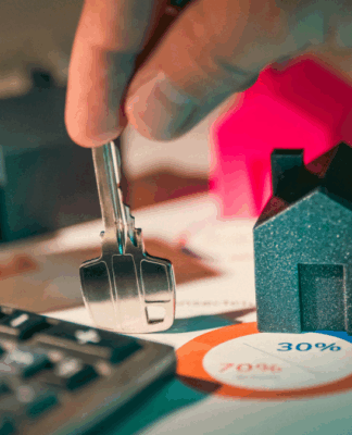 Pessoa segura uma chave sobre uma mesa com miniaturas de casas, notas de dinheiro, calculadora e gráfico circular, representando o mercado imobiliário e o custo do aluguel.