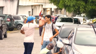 Em uma rua com carros estacionados, um homem de boné azul e camiseta clara dá um tapa no rosto de outro homem mais jovem, que usa mochila e camiseta branca com estampa geométrica. O jovem parece surpreso e inclina o rosto para o lado. Ao fundo, há outras pessoas caminhando e um homem com colete laranja acenando com o braço levantado. A cena ocorre em ambiente urbano, durante o dia