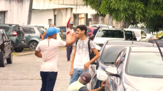 Em uma rua com carros estacionados, um homem de boné azul e camiseta clara dá um tapa no rosto de outro homem mais jovem, que usa mochila e camiseta branca com estampa geométrica. O jovem parece surpreso e inclina o rosto para o lado. Ao fundo, há outras pessoas caminhando e um homem com colete laranja acenando com o braço levantado. A cena ocorre em ambiente urbano, durante o dia