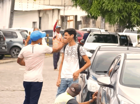 Em uma rua com carros estacionados, um homem de boné azul e camiseta clara dá um tapa no rosto de outro homem mais jovem, que usa mochila e camiseta branca com estampa geométrica. O jovem parece surpreso e inclina o rosto para o lado. Ao fundo, há outras pessoas caminhando e um homem com colete laranja acenando com o braço levantado. A cena ocorre em ambiente urbano, durante o dia