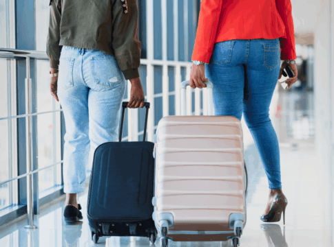 Duas mulheres caminham pelo aeroporto, cada uma puxando sua mala.