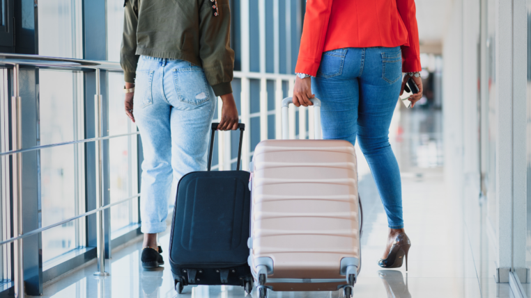 Duas mulheres caminham pelo aeroporto, cada uma puxando sua mala.