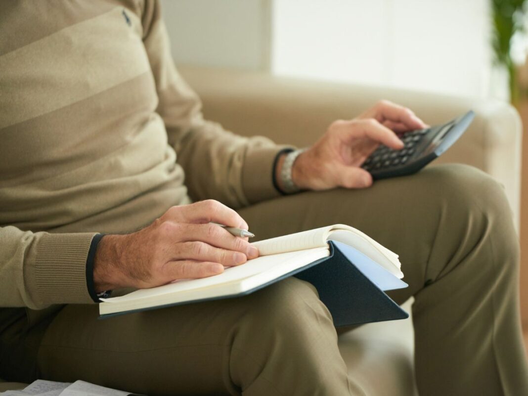Homem branco sentado segurando uma calculadora e um escrevendo em um caderno.