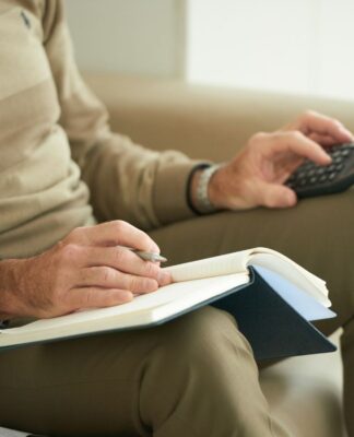 Homem branco sentado segurando uma calculadora e um escrevendo em um caderno.