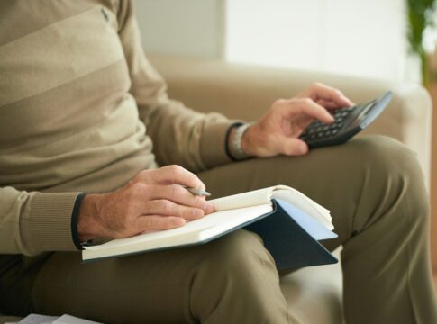 Homem branco sentado segurando uma calculadora e um escrevendo em um caderno.