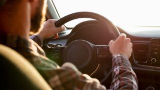 A imagem mostra um homem de costas dirigindo com uma camisa xadrez e as duas mãos no volante