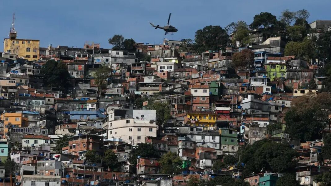 Uma foto panorâmica, tirada em plano aberto, que mostra uma grande favela ou comunidade verticalizada no Rio de Janeiro, estendendo-se por um morro. A cena é capturada em um dia claro, com céu azul.