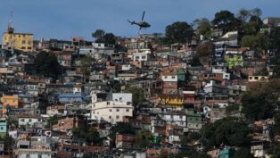 Uma foto panorâmica, tirada em plano aberto, que mostra uma grande favela ou comunidade verticalizada no Rio de Janeiro, estendendo-se por um morro. A cena é capturada em um dia claro, com céu azul.