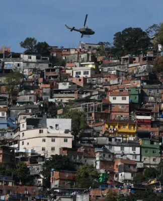 Uma foto panorâmica, tirada em plano aberto, que mostra uma grande favela ou comunidade verticalizada no Rio de Janeiro, estendendo-se por um morro. A cena é capturada em um dia claro, com céu azul.