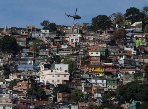 Uma foto panorâmica, tirada em plano aberto, que mostra uma grande favela ou comunidade verticalizada no Rio de Janeiro, estendendo-se por um morro. A cena é capturada em um dia claro, com céu azul.