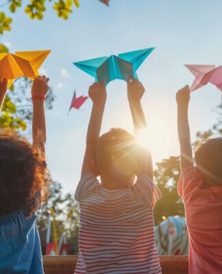 A imagem mostra três crianças brincando de costas em um parque com origamis nas mãos