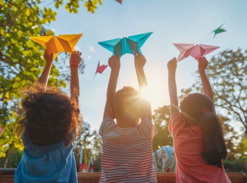 A imagem mostra três crianças brincando de costas em um parque com origamis nas mãos