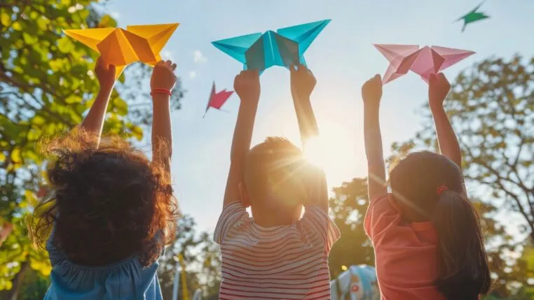 A imagem mostra três crianças brincando de costas em um parque com origamis nas mãos