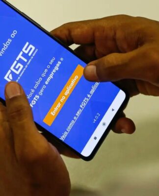 A imagem mostra as mãos de uma pessoa segurando o celular. Na tela do aparelho está o aplicativo do FGTS