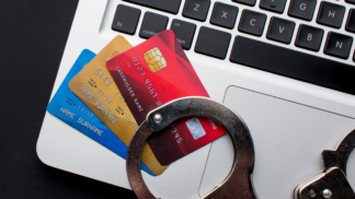 Cartões de crédito coloridos sobre o teclado de um notebook prateado, com um par de algemas apoiado por cima, simbolizando crimes financeiros e fraudes bancárias online