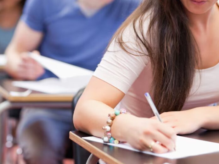 Aluna sentada em uma carteira de escola com uma caneta na mão, escrevendo em uma folha