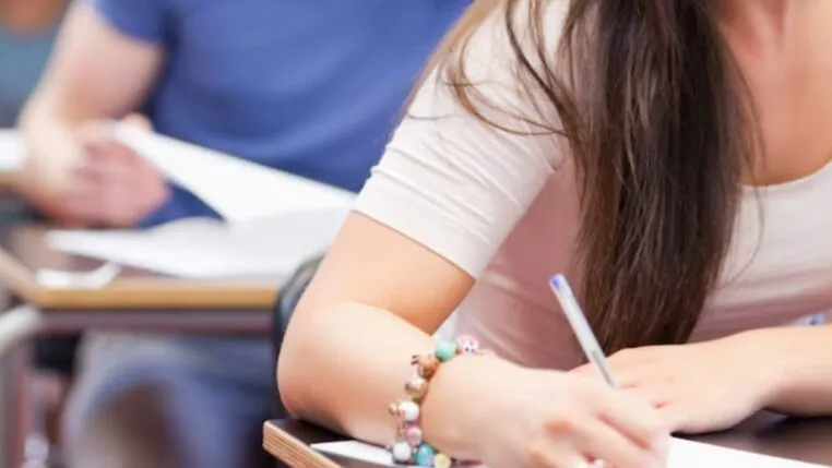 Aluna sentada em uma carteira de escola com uma caneta na mão, escrevendo em uma folha