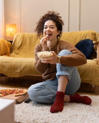 Mulher assistindo televisão em uma sala com um pote de pipoca na mão.