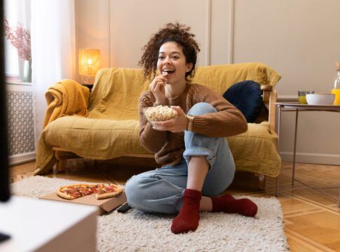 Mulher assistindo televisão em uma sala com um pote de pipoca na mão.