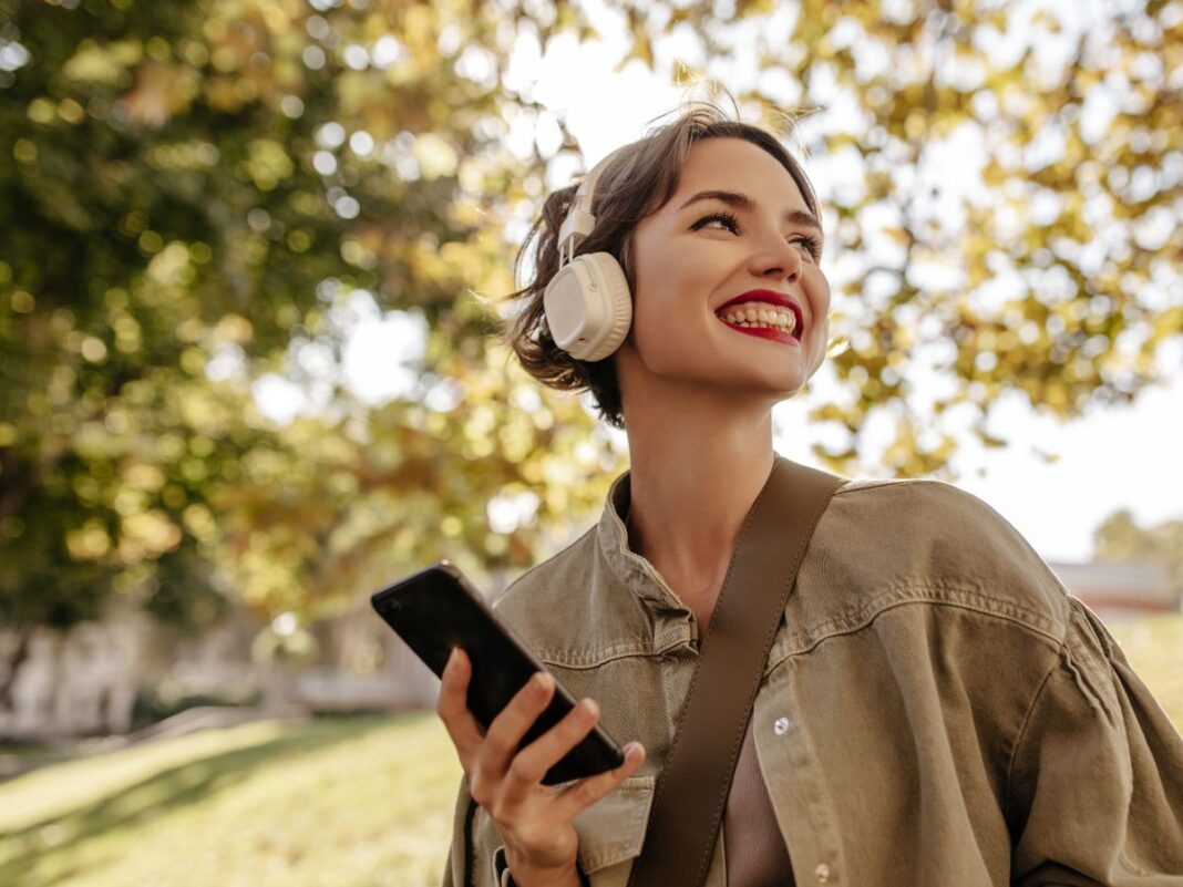 Mulher branca usando casaco e batom vermelho está com um fone de ouvido e segurando um celular em uma paisagem de árvores.