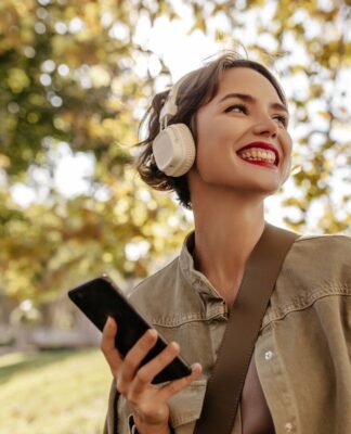 Mulher branca usando casaco e batom vermelho está com um fone de ouvido e segurando um celular em uma paisagem de árvores.