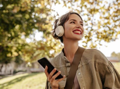Mulher branca usando casaco e batom vermelho está com um fone de ouvido e segurando um celular em uma paisagem de árvores.