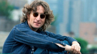 Johnn Lennon sentado de lado, em um espaço aberto e gramado, com camisa jeans e óculos de sol redondo.
