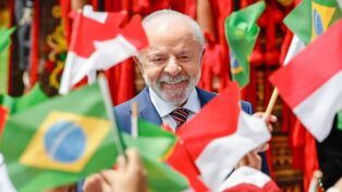 Uma foto em plano close-up registra o Presidente do Brasil, Luiz Inácio Lula da Silva, sorrindo, enquanto é cercado por bandeiras do Brasil e da Indonésia, seguradas por mãos que não aparecem por completo. As bandeiras em primeiro plano, como a brasileira à esquerda inferior e a indonésia à direita inferior, estão bem nítidas, em contraste com as do fundo.