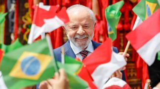 Uma foto em plano close-up registra o Presidente do Brasil, Luiz Inácio Lula da Silva, sorrindo, enquanto é cercado por bandeiras do Brasil e da Indonésia, seguradas por mãos que não aparecem por completo. As bandeiras em primeiro plano, como a brasileira à esquerda inferior e a indonésia à direita inferior, estão bem nítidas, em contraste com as do fundo.