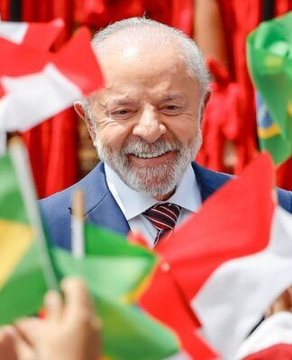 Uma foto em plano close-up registra o Presidente do Brasil, Luiz Inácio Lula da Silva, sorrindo, enquanto é cercado por bandeiras do Brasil e da Indonésia, seguradas por mãos que não aparecem por completo. As bandeiras em primeiro plano, como a brasileira à esquerda inferior e a indonésia à direita inferior, estão bem nítidas, em contraste com as do fundo.