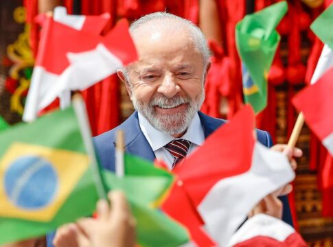 Uma foto em plano close-up registra o Presidente do Brasil, Luiz Inácio Lula da Silva, sorrindo, enquanto é cercado por bandeiras do Brasil e da Indonésia, seguradas por mãos que não aparecem por completo. As bandeiras em primeiro plano, como a brasileira à esquerda inferior e a indonésia à direita inferior, estão bem nítidas, em contraste com as do fundo.