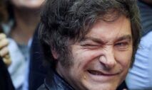 A imagem é um retrato em plano muito fechado do rosto do político argentino Javier Milei. Ele está em primeiro plano, com o rosto virado ligeiramente para a esquerda. Sua expressão é intensa e caricata: ele sorri amplamente, mostrando os dentes, e aperta o olho direito (piscando ou semicerrando) de forma exagerada, o que acentua as rugas de expressão ao redor dos olhos.