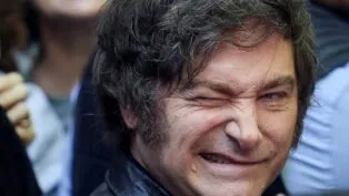 A imagem é um retrato em plano muito fechado do rosto do político argentino Javier Milei. Ele está em primeiro plano, com o rosto virado ligeiramente para a esquerda. Sua expressão é intensa e caricata: ele sorri amplamente, mostrando os dentes, e aperta o olho direito (piscando ou semicerrando) de forma exagerada, o que acentua as rugas de expressão ao redor dos olhos.