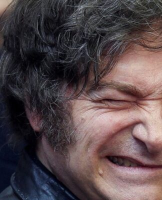 A imagem é um retrato em plano muito fechado do rosto do político argentino Javier Milei. Ele está em primeiro plano, com o rosto virado ligeiramente para a esquerda. Sua expressão é intensa e caricata: ele sorri amplamente, mostrando os dentes, e aperta o olho direito (piscando ou semicerrando) de forma exagerada, o que acentua as rugas de expressão ao redor dos olhos.