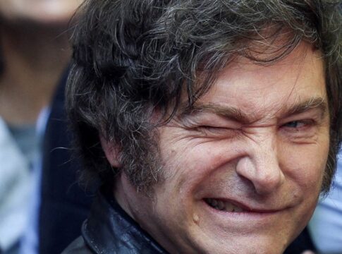 A imagem é um retrato em plano muito fechado do rosto do político argentino Javier Milei. Ele está em primeiro plano, com o rosto virado ligeiramente para a esquerda. Sua expressão é intensa e caricata: ele sorri amplamente, mostrando os dentes, e aperta o olho direito (piscando ou semicerrando) de forma exagerada, o que acentua as rugas de expressão ao redor dos olhos.