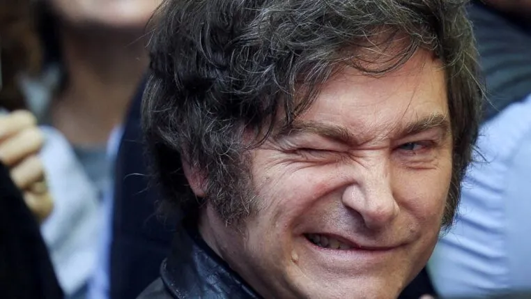 A imagem é um retrato em plano muito fechado do rosto do político argentino Javier Milei. Ele está em primeiro plano, com o rosto virado ligeiramente para a esquerda. Sua expressão é intensa e caricata: ele sorri amplamente, mostrando os dentes, e aperta o olho direito (piscando ou semicerrando) de forma exagerada, o que acentua as rugas de expressão ao redor dos olhos.
