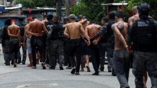 A fotografia, capturada em um ambiente de rua, numa comunidade, mostra um grupo de homens, alguns deles detidos, sendo conduzidos por policiais militares.