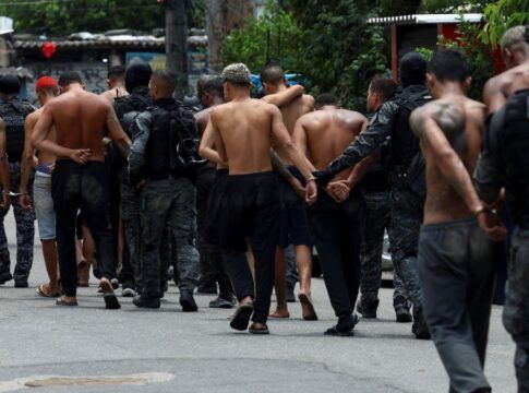 A fotografia, capturada em um ambiente de rua, numa comunidade, mostra um grupo de homens, alguns deles detidos, sendo conduzidos por policiais militares.