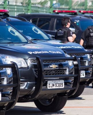Viaturas estão enfileiradas num estacionamento. Ao lado dos veículos policiais, aparecem três policiais civis.