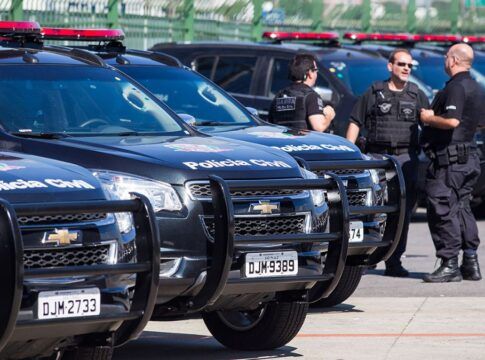 Viaturas estão enfileiradas num estacionamento. Ao lado dos veículos policiais, aparecem três policiais civis.