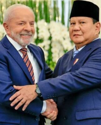 Uma foto em plano médio e em ambiente interno registra o aperto de mãos entre o Presidente do Brasil, Luiz Inácio Lula da Silva (à esquerda), e o Presidente da Indonésia, Prabowo Subianto (à direita), durante um encontro oficial, em Jacarta. No fundo, notam-se as bandeiras nacionais, ligeiramente desfocadas: a bandeira da Indonésia (vermelha e branca) e a bandeira do Brasil (verde, amarela e azul), confirmando o contexto diplomático da reunião. A iluminação é forte, de ambiente formal, com decorações florais brancas visíveis atrás dos líderes.