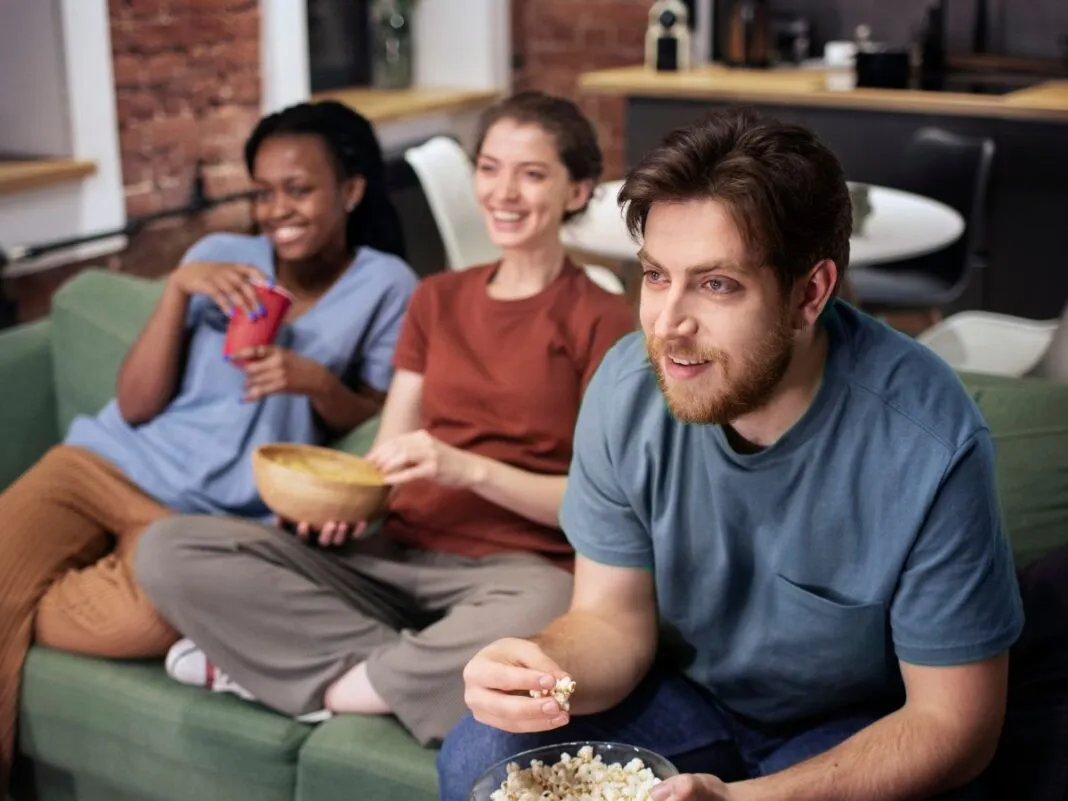 Um homem e duas mulheres estão sentados em um sofá com pipoca na mão e olhando para uma TV