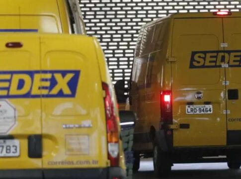 Imagem mostra a traseira de duas vans de entrega dos Correios, ambas na cor amarela característica, em uma área de depósito ou doca de carregamento com uma parede de metal perfurada ao fundo.