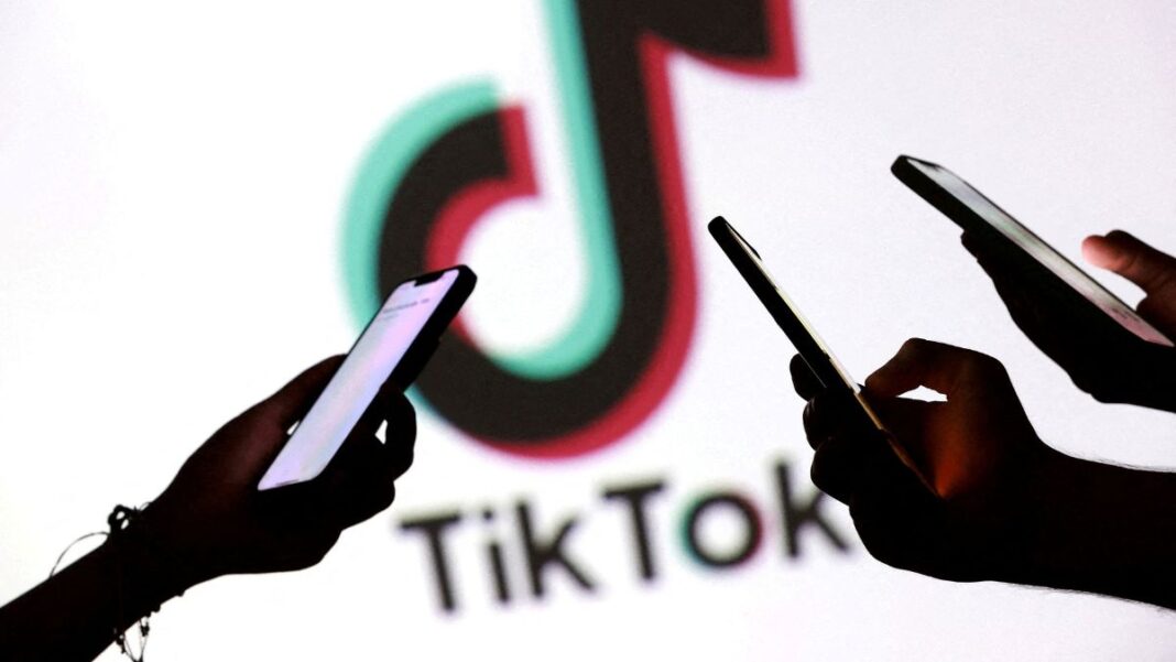 Imagem mostra as mãos de três pessoas mexendo no celular, em primeiro plano. No segundo plano, há o logo da empresa TikTok.